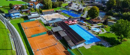 Luftaufnahme eines Sportkomplexes mit Tennisplätzen, einem Schwimmbad und einem Volleyballfeld. Gebäude und eine Brücke sind in der Ferne sichtbar.