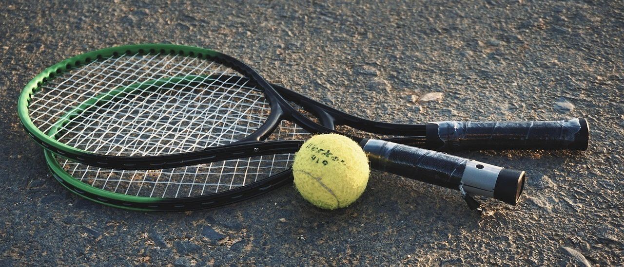 Ein Tennisschläger und ein Tennisball mit der Marke Wilson liegen auf dem Boden. Der Schläger ist schwarz mit grünem Rand. Der Ball ist gelb mit etwas Schrift darauf.