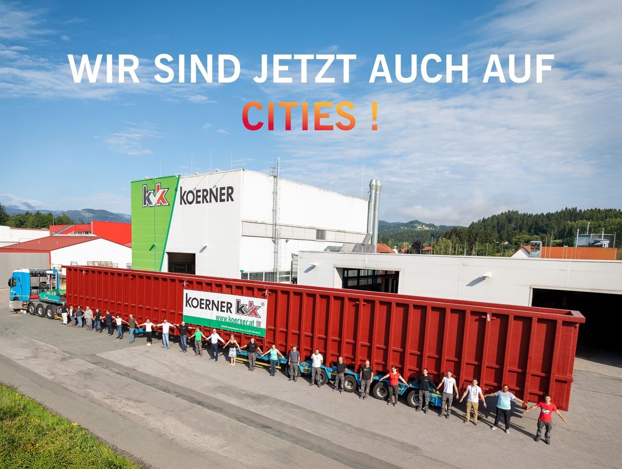 Eine Gruppe von Menschen steht vor einem roten Container mit dem Text Koerner darauf. Dahinter befindet sich ein großes Gebäude mit dem Text Koerner an der Seite. Der Himmel ist blau mit Wolken.