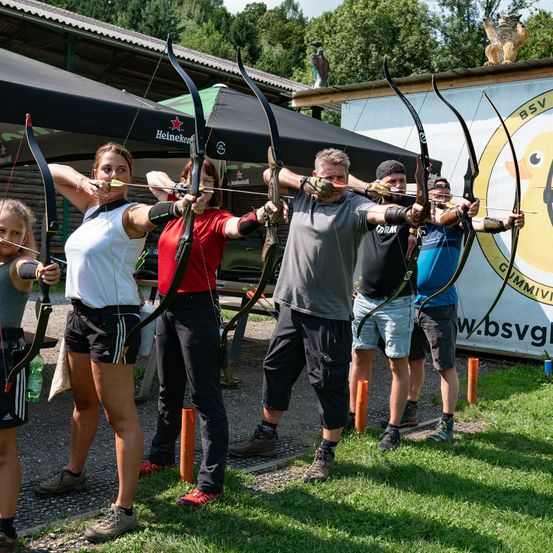 Eine Gruppe von Personen in Sportkleidung, einschließlich Handschuhen, nimmt an einer Bogenschießveranstaltung auf einem Grasfeld teil. Sie halten Bögen und Pfeile, und einige tragen Mützen. Im Hintergrund befinden sich Zelte, eine Wand mit einem gelben Entenlogo und eine Bank.