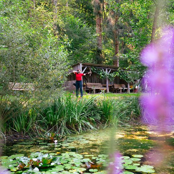 Eine Person steht in einem üppigen Garten und hält einen Bogen. Hinter ihr ist eine rustikale Holzhütte mit Bänken zu sehen. Das Gebiet ist von viel Grün umgeben, und ein Teich mit Seerosen liegt vor ihr.