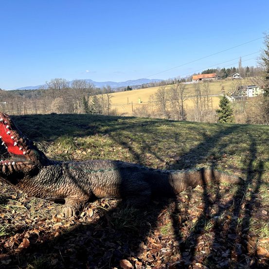 Eine lebensgroße, bunte Dinosaurierstatue liegt auf dem Boden, der Kopf gehoben mit offenen Kiefern. Der Hintergrund ist ein Grasfeld mit Bäumen und einem entfernten Haus. Berge und Stromleitungen sind im Hintergrund zu sehen.