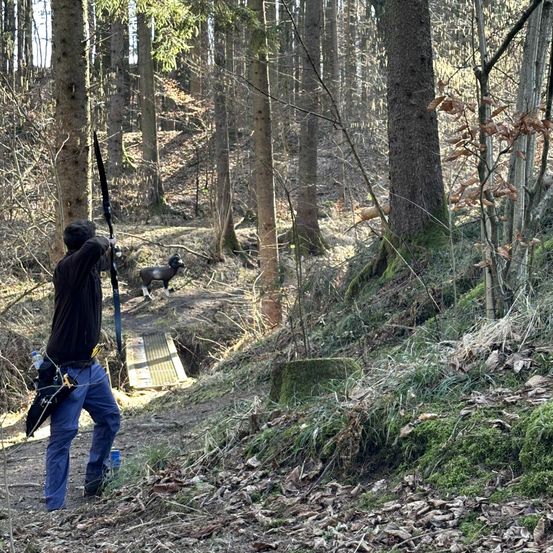 Ein Mann steht in einem Wald, einen Bogen haltend, mit einem Schaf im Hintergrund. Der Boden ist mit abgefallenen Blättern und Moos bedeckt.