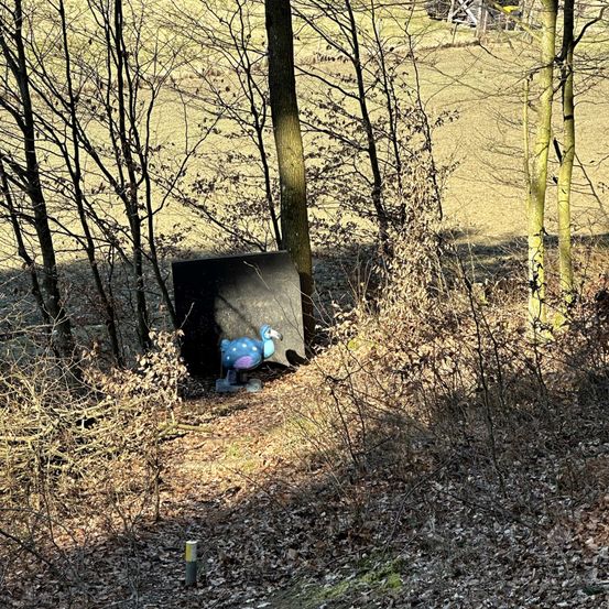 Eine leuchtend blaue Vogelstatue steht neben einem schwarzen Objekt in einem laubbedeckten Wald. Der Wald hat kahle Bäume und im Hintergrund ein weites, grünes Feld.