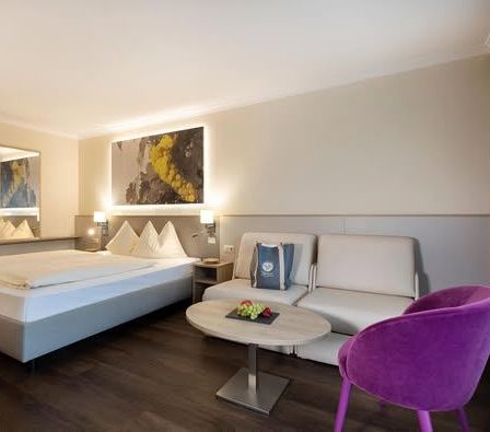 Ein Hotelzimmer mit einem Bett, einer weißen Couch, einem lila Stuhl und einem Tisch mit einer Tasche darauf. Das Zimmer hat einen Spiegel und ein Gemälde an der Wand.