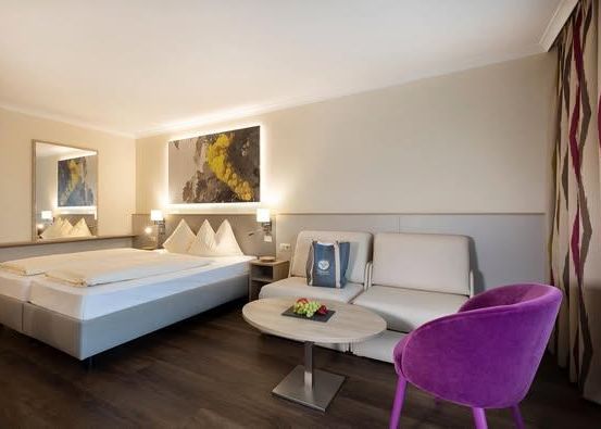 Ein Hotelzimmer mit einem Bett, einer weißen Couch, einem lila Stuhl und einem Tisch mit einer Tasche darauf. Das Zimmer hat einen Spiegel und ein Gemälde an der Wand.