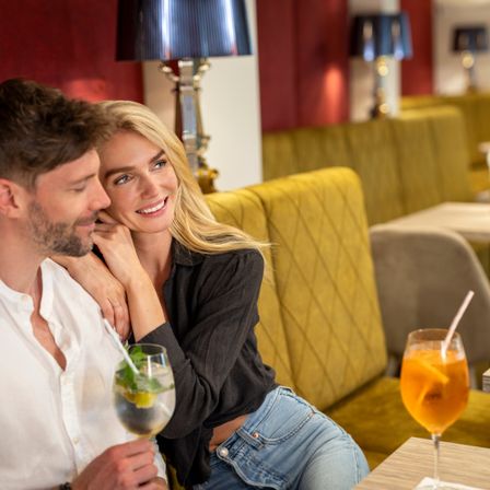 Ein Paar in einem Restaurant, der Mann und die Frau lächeln und halten Getränke. Sie sitzen auf einer gelben Couch, und die Frau lehnt sich an die Schulter des Mannes.
