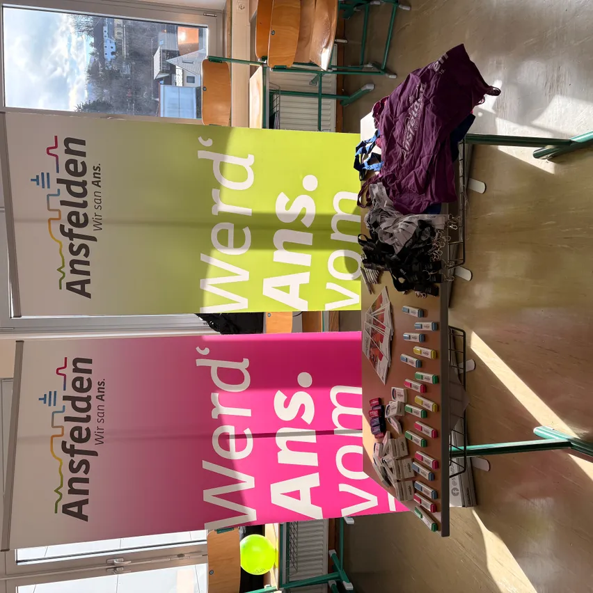 Eine Ausstellung in einem Raum mit zwei Bannern und einem Tisch mit einem gelben Luftballon und verschiedenen Gegenständen. Ein Blick auf einen bewölkten Himmel ist durch das Fenster sichtbar.