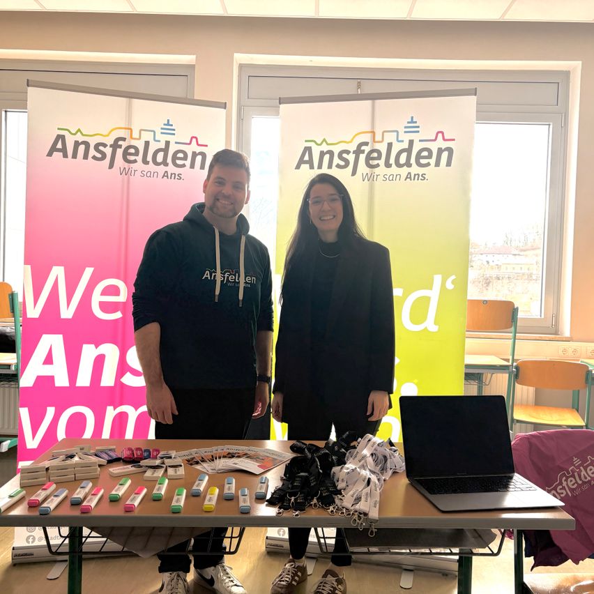 Zwei Personen stehen vor einem Tisch mit verschiedenen Gegenständen. Ein Mann trägt eine Kapuzenjacke und eine Frau hat eine Brille. Sie befinden sich in einem Raum mit Bannern, die Ansfelden lesen.