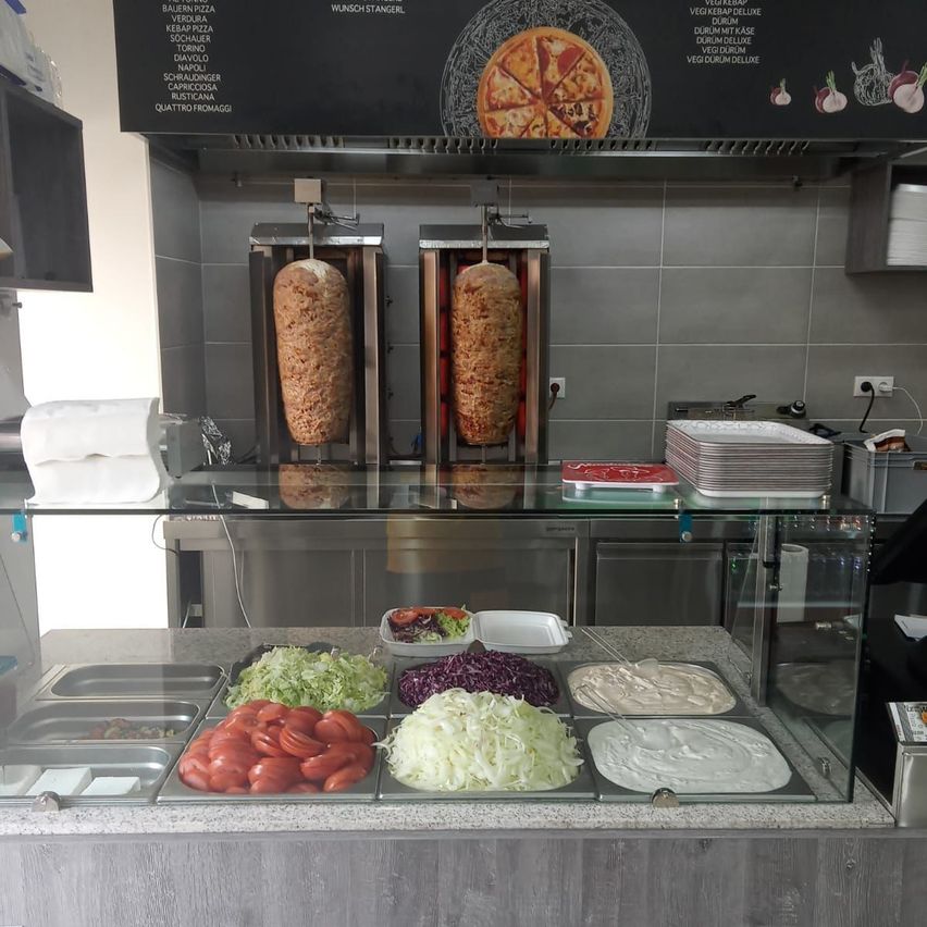 Eine Restaurantküche mit einer Glasvitrine, die verschiedene Zutaten wie Tomaten, Kohl und Saucen enthält. Im Hintergrund werden zwei Fleischspieße geröstet.