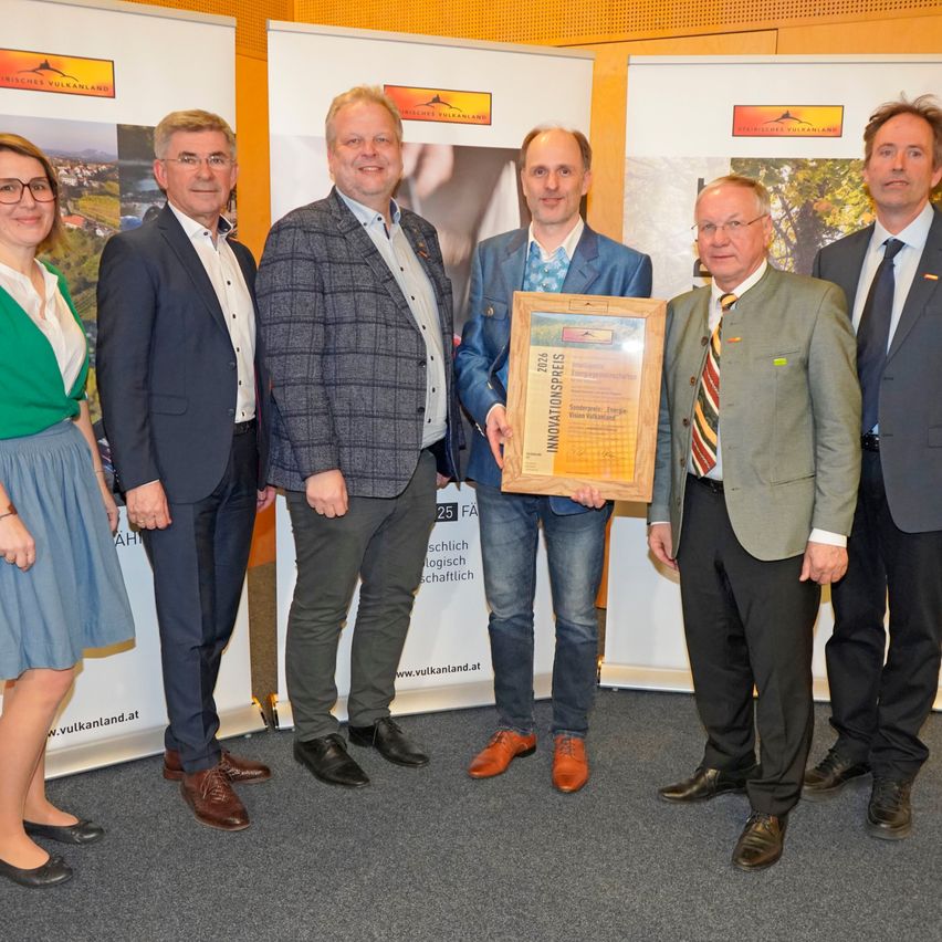 Sechs Menschen in formeller Kleidung stehen in einer Reihe und posieren für ein Foto. Einer von ihnen hält eine gerahmte Auszeichnungsurkunde. Der Hintergrund zeigt Werbebanner.