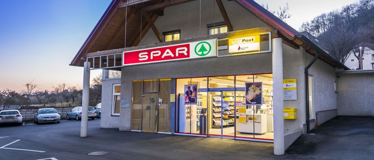 Die Vorderseite eines Spar-Geschäfts mit einem Postamt im Inneren. Das Gebäude hat ein dreieckiges Dach, Glastüren und Fenster. Der Laden hat ein gelbes Schild mit dem Wort 'Post' darauf.