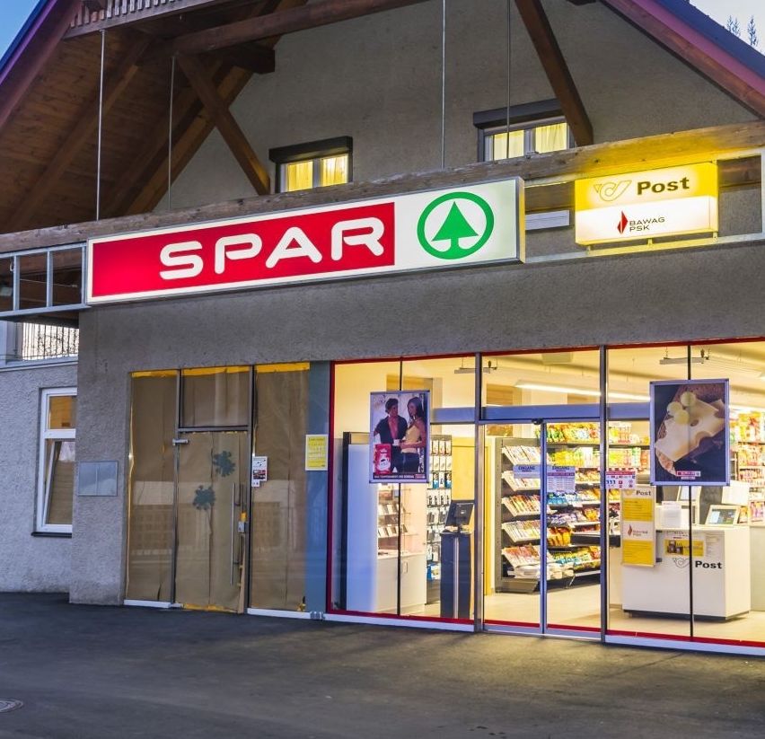 Die Vorderseite eines Spar-Geschäfts mit einem Postamt im Inneren. Das Gebäude hat ein dreieckiges Dach, Glastüren und Fenster. Der Laden hat ein gelbes Schild mit dem Wort 'Post' darauf.