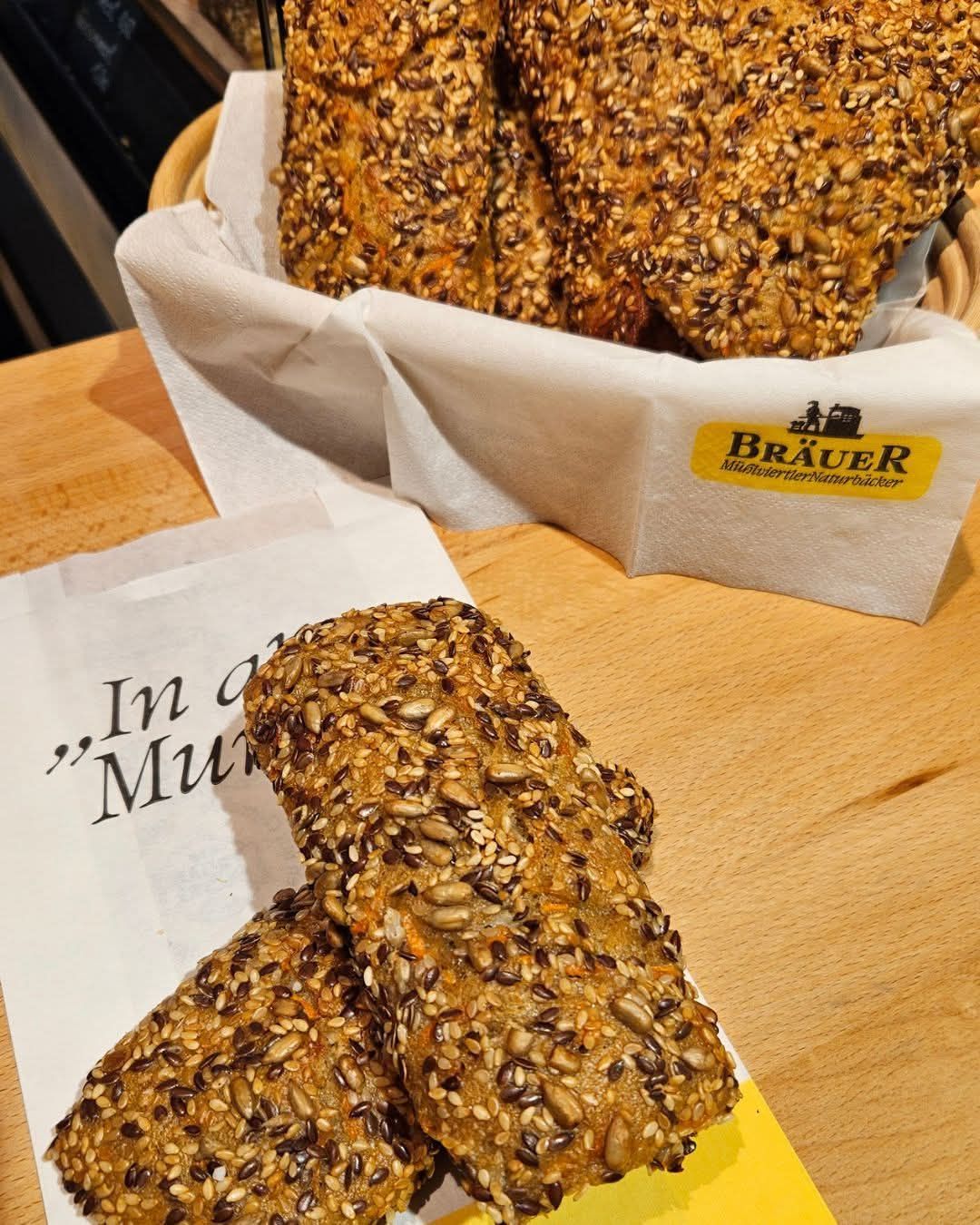 A close-up of a basket of whole grain bread with sesame seeds on a wooden table. The basket has a white paper lining with the word 'Bräuer' on it.