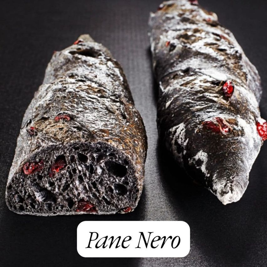 Zwei dunkel gefärbte Laibe mit sichtbaren roten Cranberries, beschriftet mit 'Pane Nero', sind auf einer schwarzen Unterlage zu sehen.
