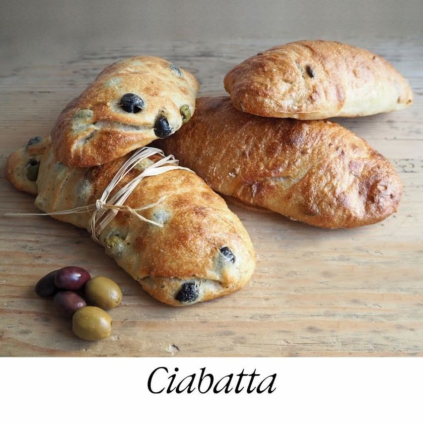 Drei Ciabatta-Brotlaibe mit Oliven, Blauschimmelkäse und grünen Oliven auf einer Holzoberfläche mit dem Wort Ciabatta in weißer Schrift.