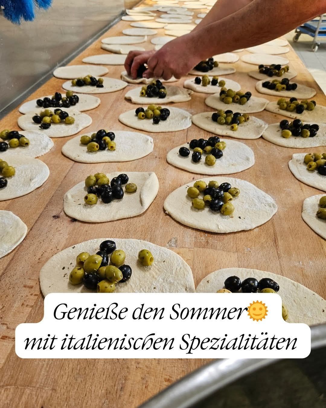 Teigplatten mit schwarzen und grünen Oliven auf einem Holztisch. Die Bildunterschrift lautet: 'Genieße den Sommer mit italienischen Spezialitäten'.