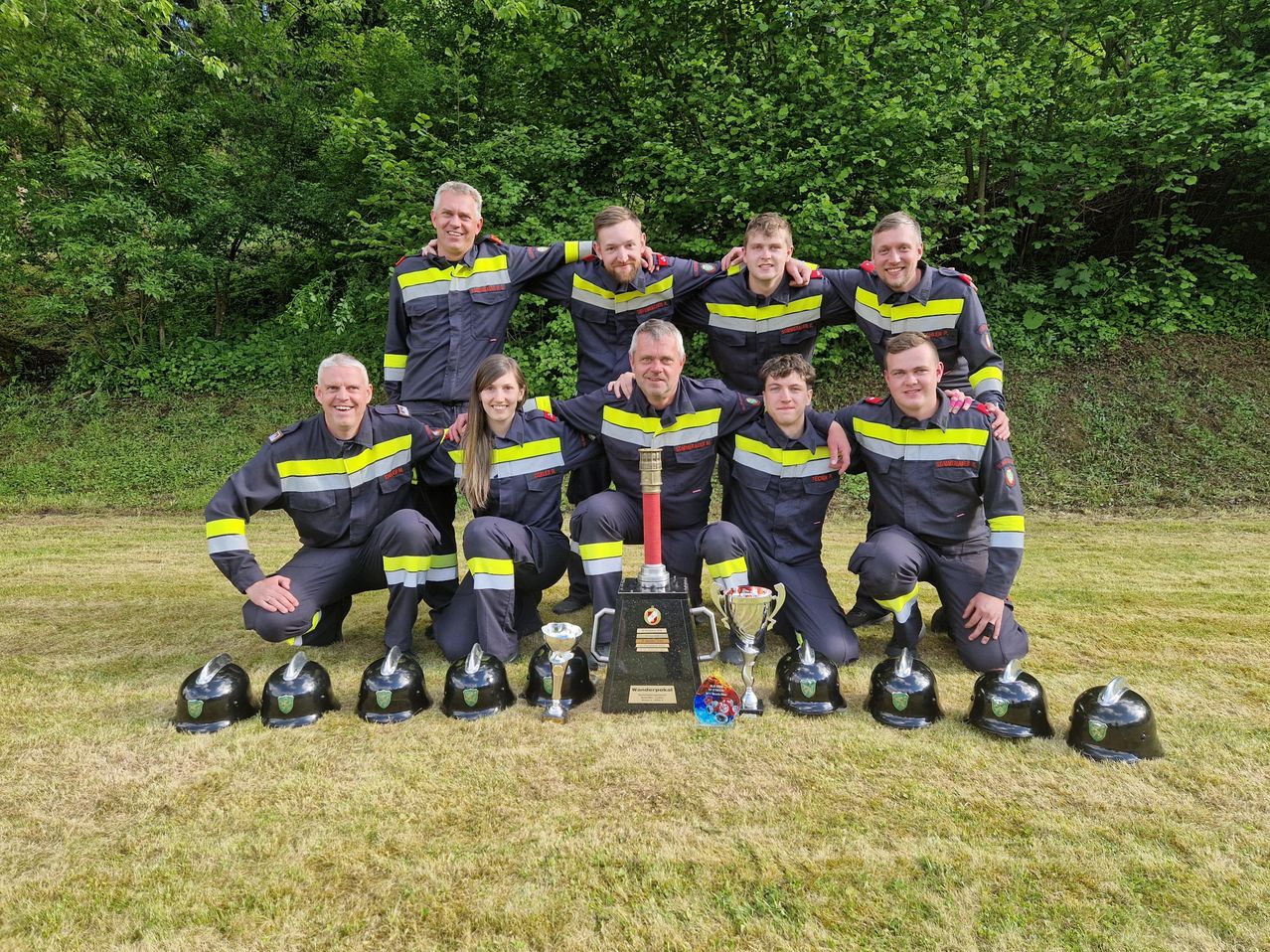 Eine Gruppe von Feuerwehrleuten posiert für ein Foto mit Helmen und Trophäen vor einem grünen Hintergrund.