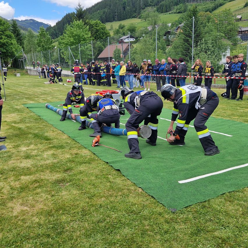 Feuerwehrleute trainieren auf einer grünen Matte, mit Zuschauern hinter Absperrungen. Sie verwenden Werkzeuge, um an Rohren zu arbeiten. Berge und Bäume sind im Hintergrund.