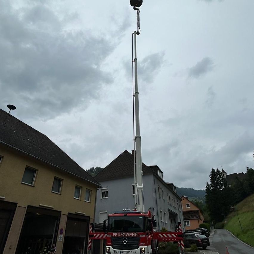 Ein Feuerwehrauto mit ausgefahrener Leiter steht vor einem Gebäude. Der Himmel ist bewölkt. Es gibt Häuser und Bäume an den Seiten.