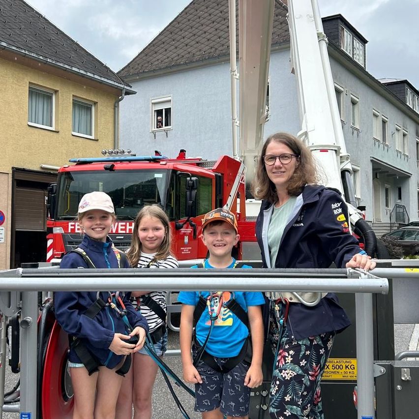 Drei Kinder und eine Frau posieren für ein Foto vor einem Feuerwehrwagen. Die Frau trägt eine Brille und eine Jacke. Die Kinder tragen Mützen und halten an Gurten fest. Ein Gebäude mit Fenstern und ein geparktes Auto sind dahinter.