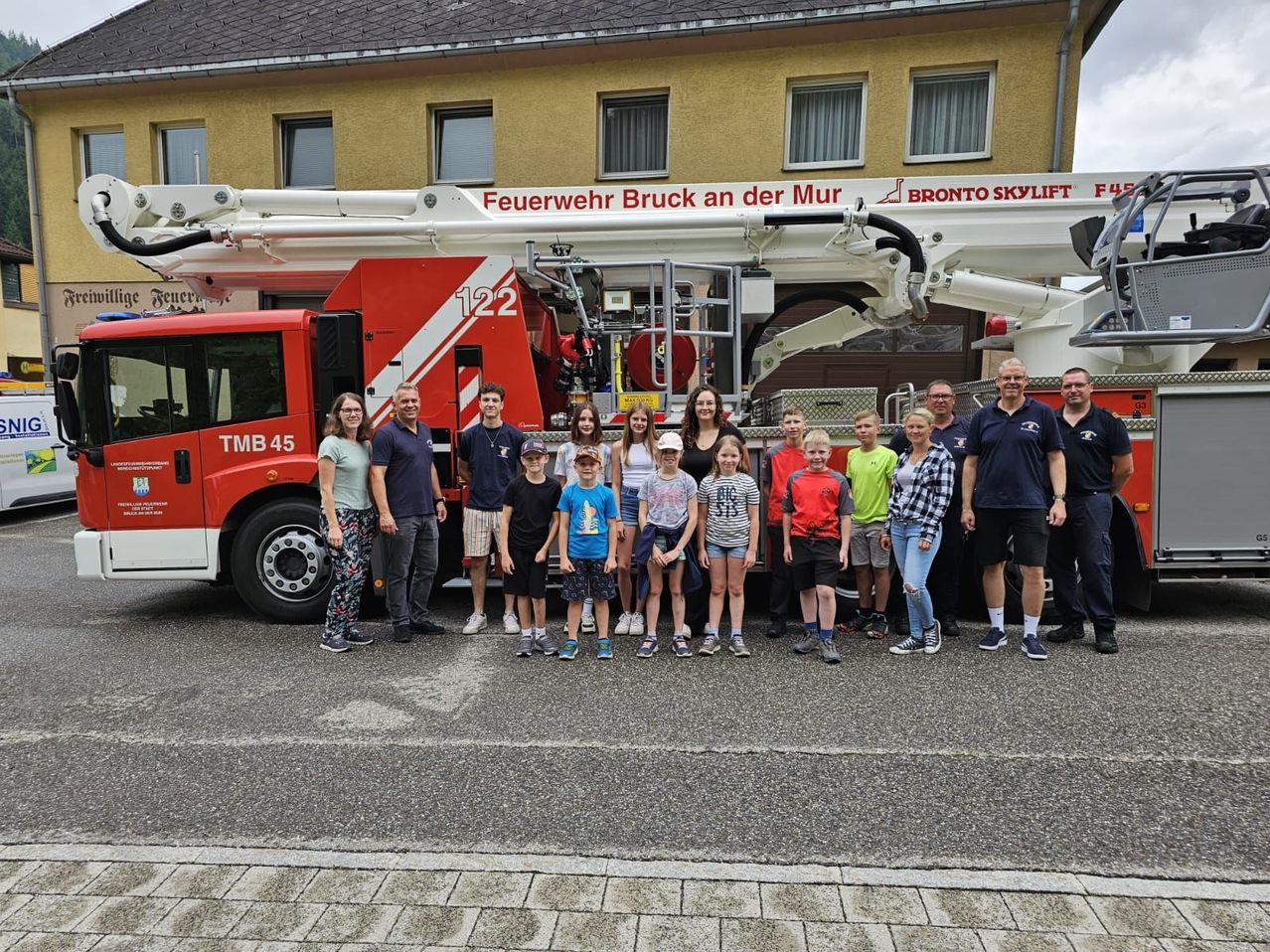 Eine Gruppe von Menschen, darunter Erwachsene und Kinder, steht vor einem roten Feuerwehrauto mit der Nummer 122. Hinter dem Auto befindet sich ein Gebäude mit den Worten Feuerwehr Bruck an der Mur und Bronto Skylift.