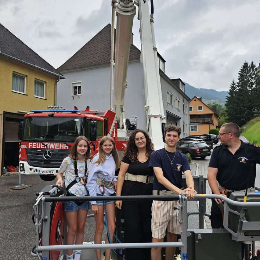 Fünf Personen stehen auf der Plattform eines Feuerwehrautos in einer Wohngegend. Hinter ihnen ist ein rotes Feuerwehrauto geparkt, und ein Haus mit Balkon ist sichtbar. Ein Berg ist in der Ferne.