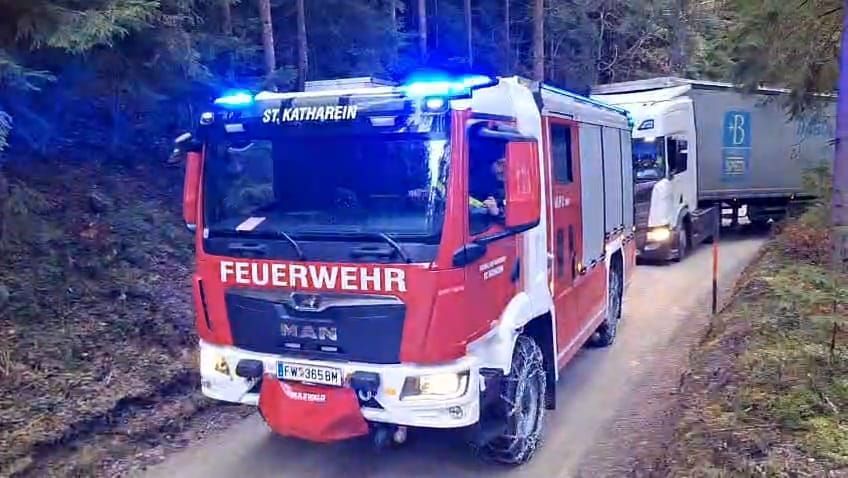 Ein roter Feuerwehrwagen mit blauen Lichtern auf dem Dach fährt eine Straße hinunter, gefolgt von einem weißen Lkw. Die Straße ist von Bäumen umgeben.