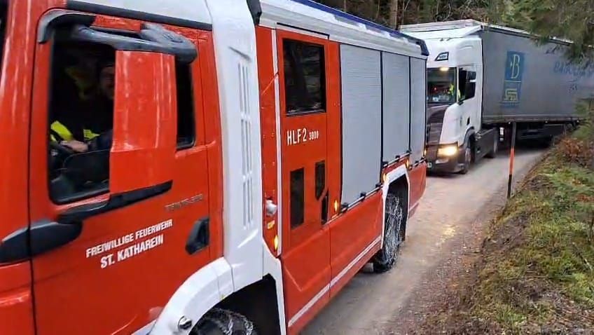 Ein roter Feuerwehrwagen mit 'HLP2 3808' auf der Tür fährt auf einer Straße, gefolgt von einem weißen LKW mit 'B' an der Seite.