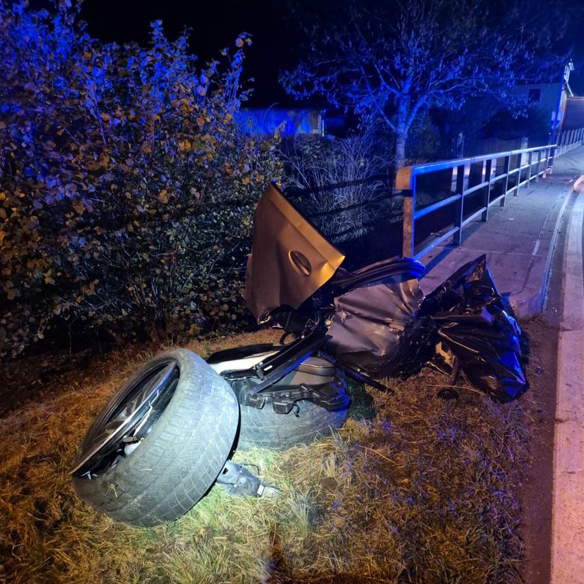 Ein beschädigtes Fahrzeug mit einem Reifen am Straßenrand, in der Nähe einer Brücke und Grünflächen unter blauer Beleuchtung bei Nacht.