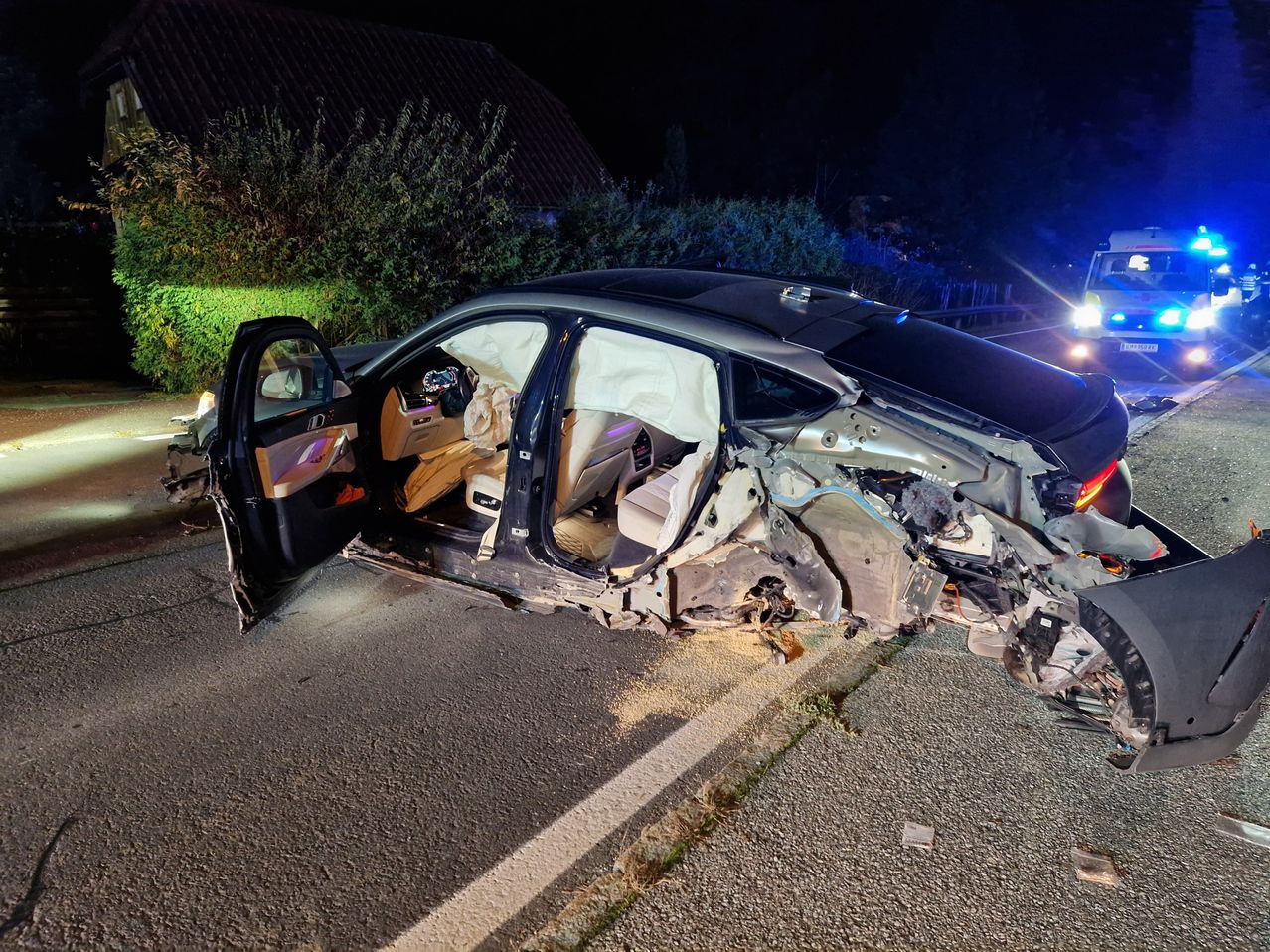 Schwer beschädigtes Auto am Straßenrand bei Nacht, mit offenen Türen und blinkenden Notfallleuchten.