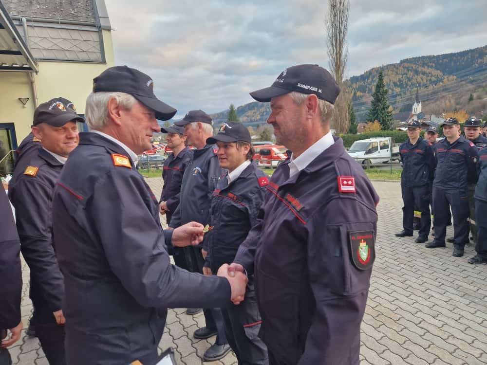 Two firefighters in uniform are shaking hands. Several other firefighters stand behind them. There are vehicles and trees in the background.