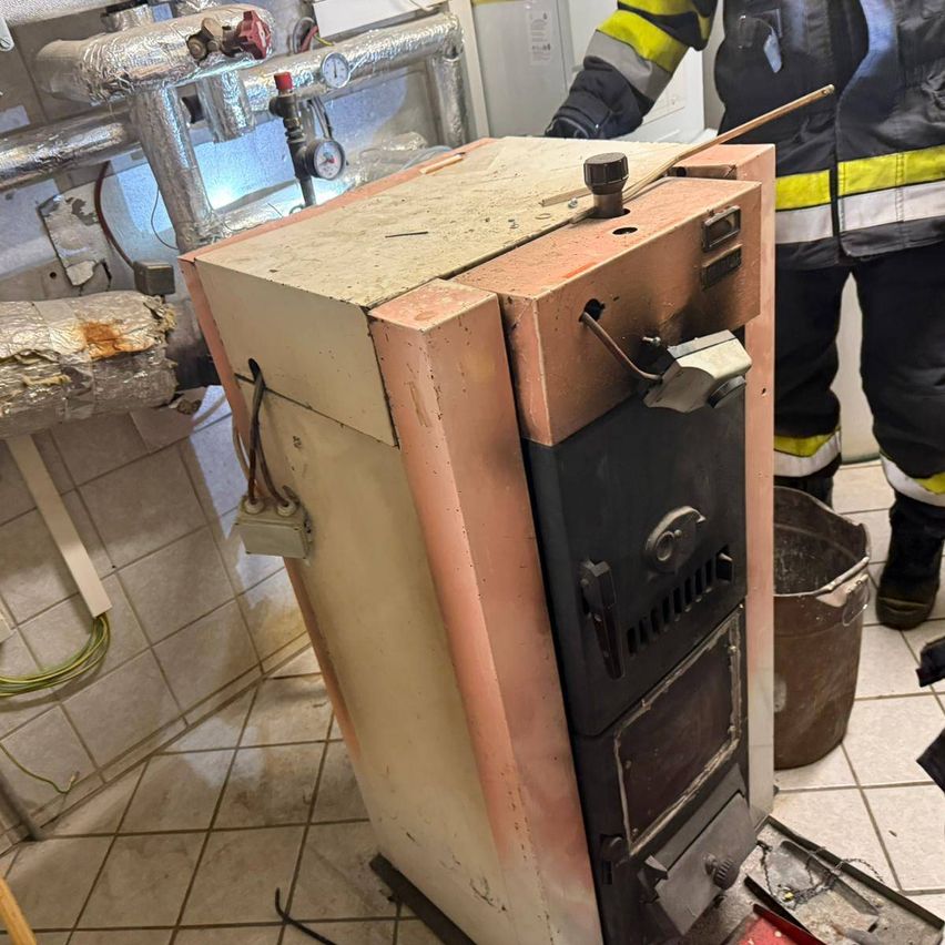 Ein alter, beschädigter Heizkörper in einem gefliesten Raum, mit einem Feuerwehrmann in der Nähe und einem Eimer auf dem Boden.