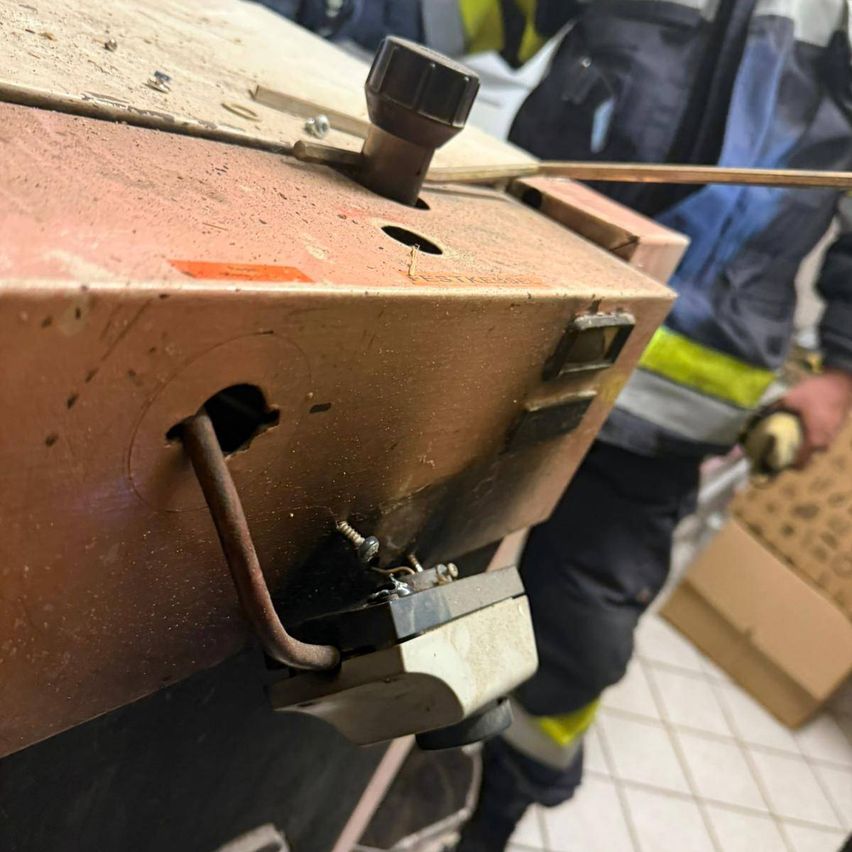 Eine Nahaufnahme eines mechanischen Geräts zeigt ein rostiges Metallstück mit einem gebogenen Metallstab und einer schwarzen Kappe. Eine Person in einem Schutzanzug steht in der Nähe.