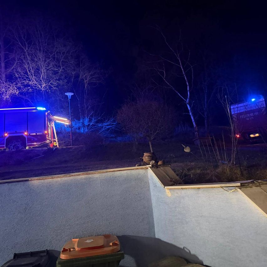 In der Nacht ist ein Rettungsfahrzeug mit blauen blinkenden Lichtern auf einem Hügel geparkt. Bäume und eine Straßenlampe sind im Hintergrund zu sehen.