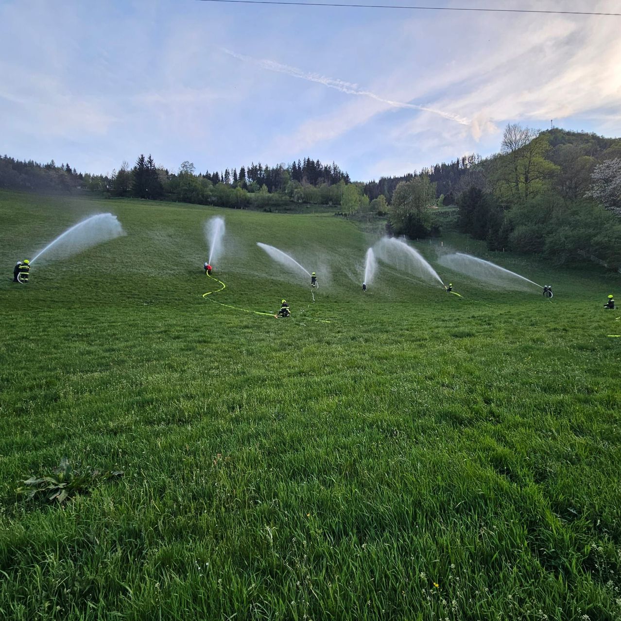 Eine Gruppe von Personen in Schutzkleidung verwendet Wassersprinkler auf einer Wiese unter einem teilweise bewölkten Himmel.