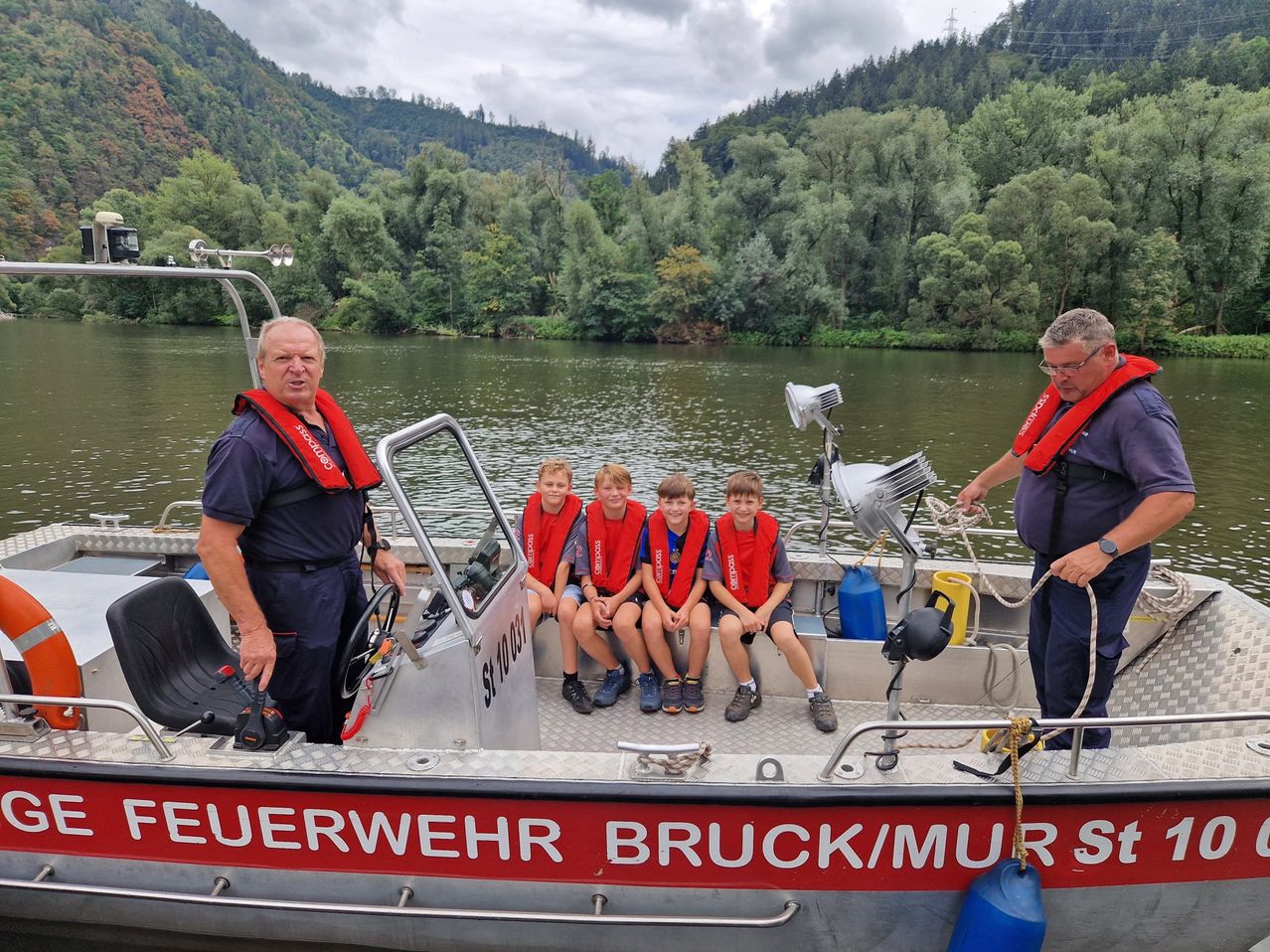 Eine Gruppe junger Jungen in Rettungswesten sitzt auf einem Boot mit zwei Männern, einer am Steuer und der andere hinten. Das Boot ist auf einem Fluss mit Bergen im Hintergrund.