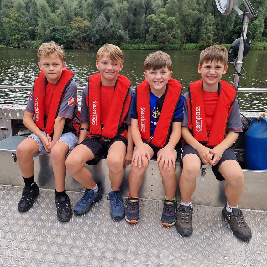 Vier Jungen in roten Schwimmwesten sitzen auf einem Boot und lächeln für ein Foto. Das Boot befindet sich auf einer ruhigen Wasserfläche mit Bäumen im Hintergrund.