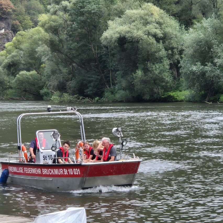 Ein rotes und silbernes Boot mit Menschen, einschließlich eines Mannes vorne, bewegt sich auf einem Fluss. Das Boot hat den Text 'Freiwillige Feuerwehr Bruckmür St 10 031' an der Seite.