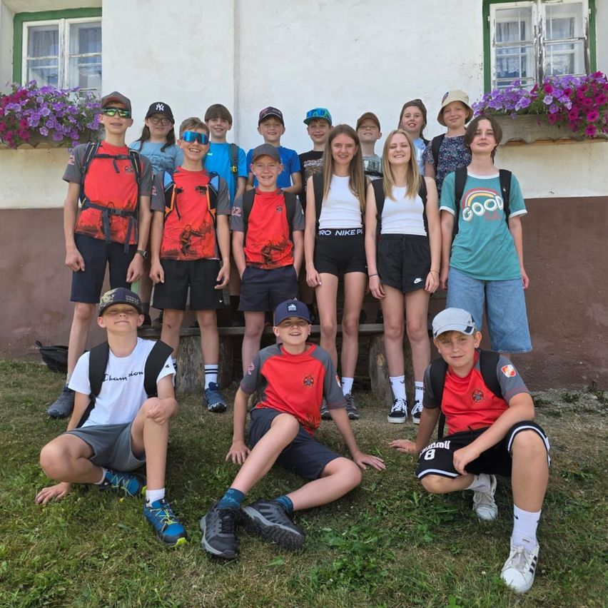 A group of children and teenagers wearing various sports gear and casual clothing pose for a photo in front of a building with flower boxes.