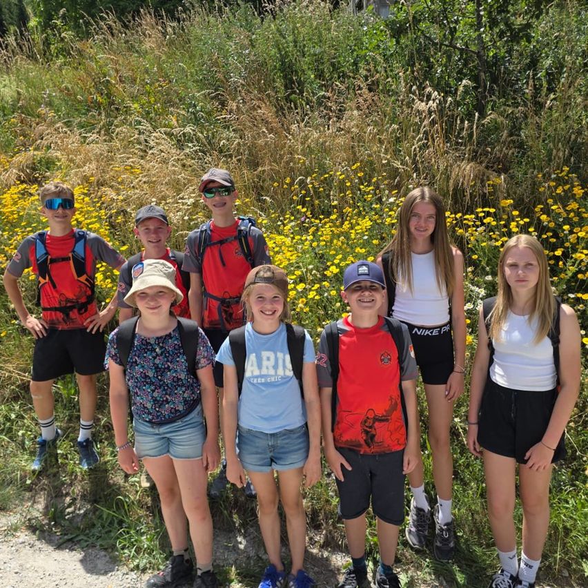 A group of young people stand together in a field of yellow flowers, wearing backpacks and hats. They are smiling and appear to be on a school trip.