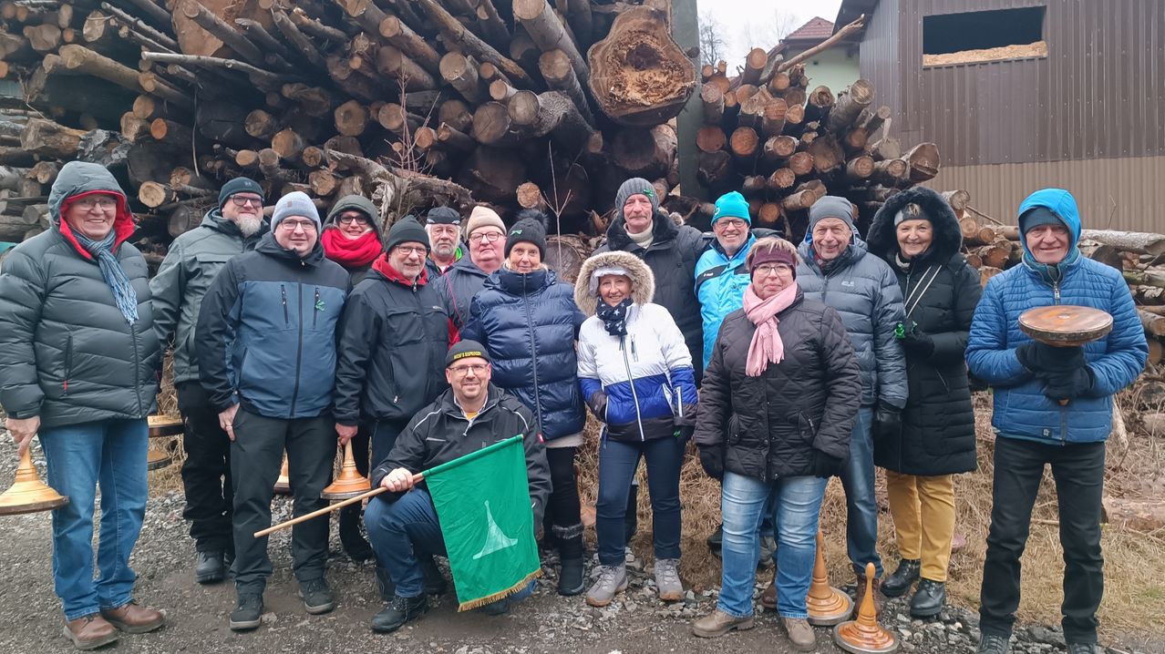 Eine Gruppe von Menschen steht vor einem großen Haufen Holzscheite. Sie sind in Winterkleidung gekleidet und einige tragen Mützen. Eine Person hält eine grüne Flagge.