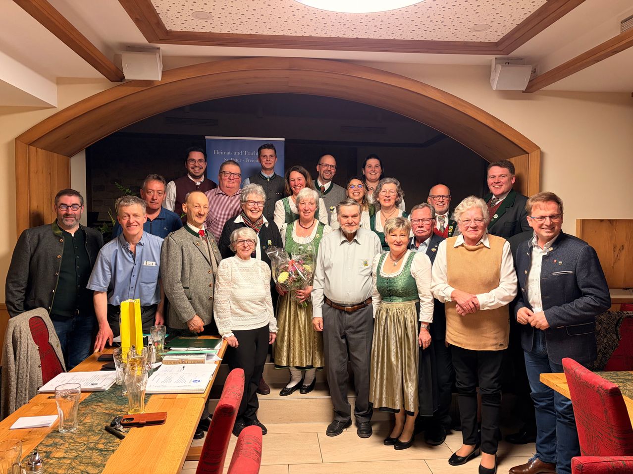 Eine Gruppe von Menschen in traditioneller Kleidung posiert für ein Foto in einem Raum mit Holzakzenten, einem Tisch und einem Banner im Hintergrund.