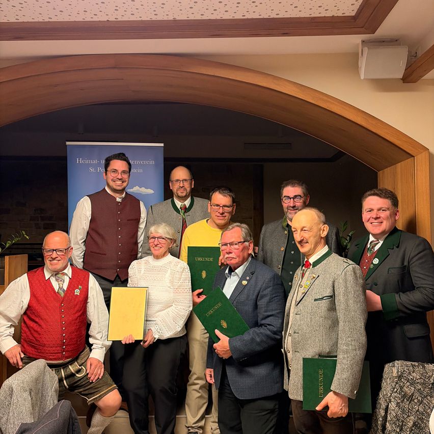 Eine Gruppe von Menschen in traditioneller Kleidung steht zusammen und posiert für ein Foto. Sie halten Urkunden in den Händen und lächeln für die Kamera. Der Ort ist drinnen mit einem hölzernen Bogen und einem Banner hinter ihnen.