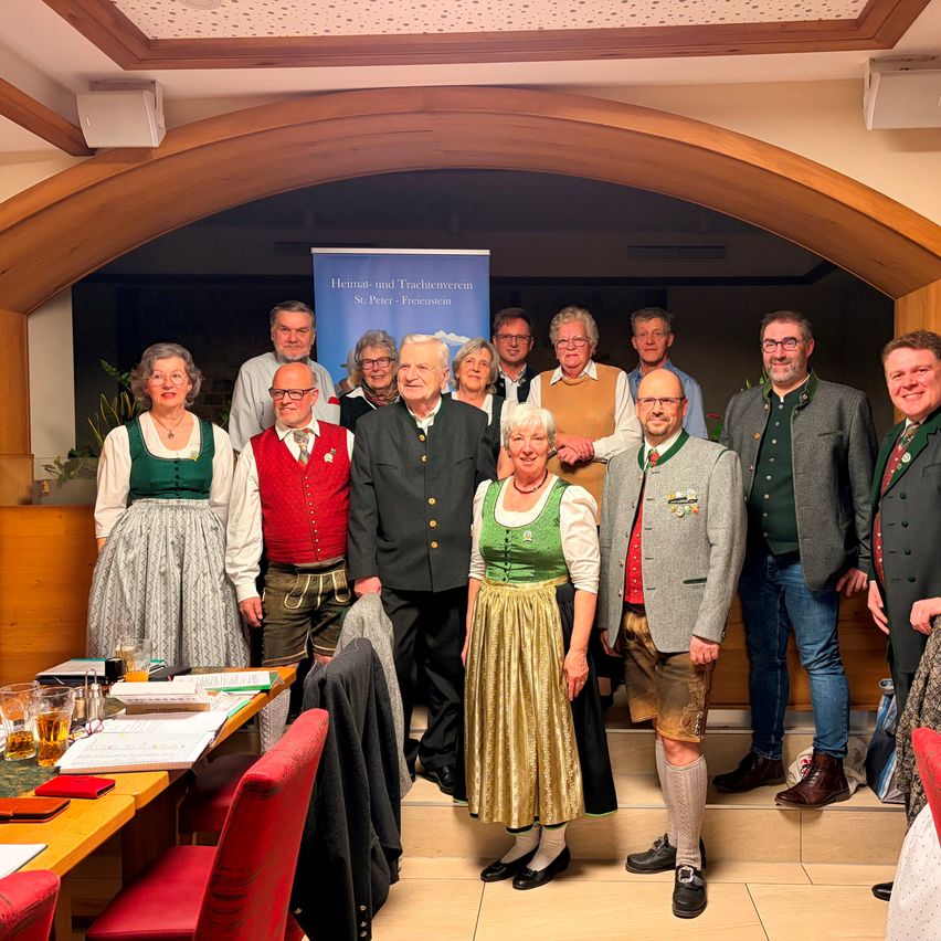 Eine Gruppe von Menschen in traditioneller Kleidung posiert für ein Foto in einem Saal, mit einem Banner und einem Tisch im Hintergrund.