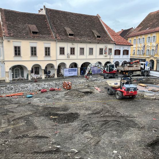 Eine Baustelle in einer Stadt mit einem großen Gebäude, einigen Fahrzeugen und einem Banner an der Wand.