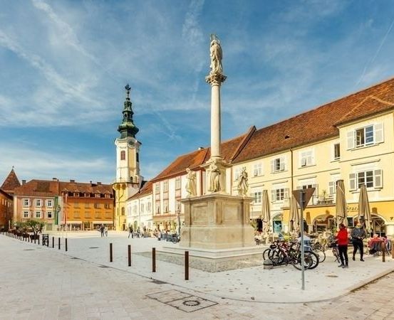 Ein Stadtplatz mit einem Denkmal in der Mitte und geparkten Fahrrädern an der Seite. Mehrere Menschen gehen herum und einige sitzen unter Sonnenschirmen in Cafés.