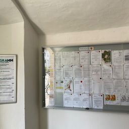 Ein Büro-Bulletin-Board mit verschiedenen angehefteten Dokumenten. Ein Schild mit grünem Text auf der linken Seite sagt Gramm. Das Board hat viele angeheftete Papiere, einige mit Logos.