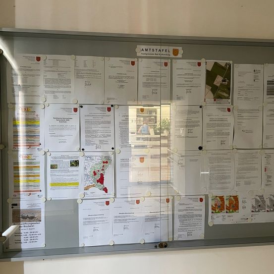 Ein Bulletinbrett mit mehreren weißen Papieren, die daran geheftet sind. Es hat ein Logo und Text auf Deutsch. Die Papiere sind mit weißen kreisförmigen Aufklebern geheftet. Es gibt eine Reflexion eines Mannes in einem blauen Hemd und einer Topfpflanze.