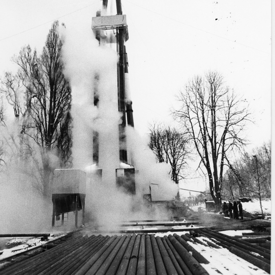 Ein Schwarz-Weiß-Foto einer Bohranlage, die Dampf abgibt, umgeben von Bäumen und Schnee, mit Menschen in der Nähe.